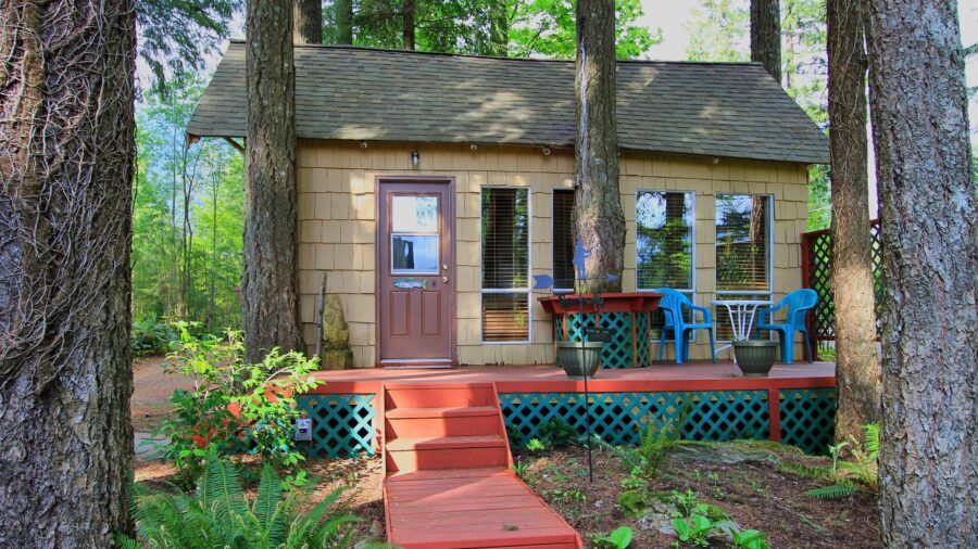 Exterior view of property painted light tan with brown door, red deck, patio table and chairs, and surrounded by tall trees and green vegetation