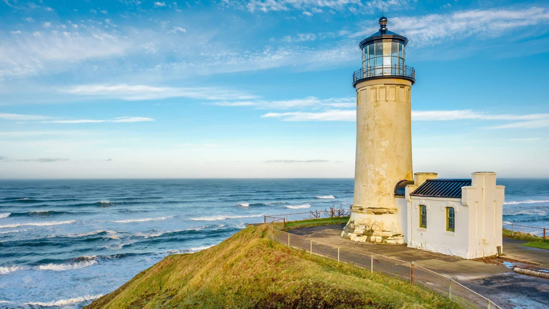 White lighthouse on a hill full of green grass looking over waves on the water