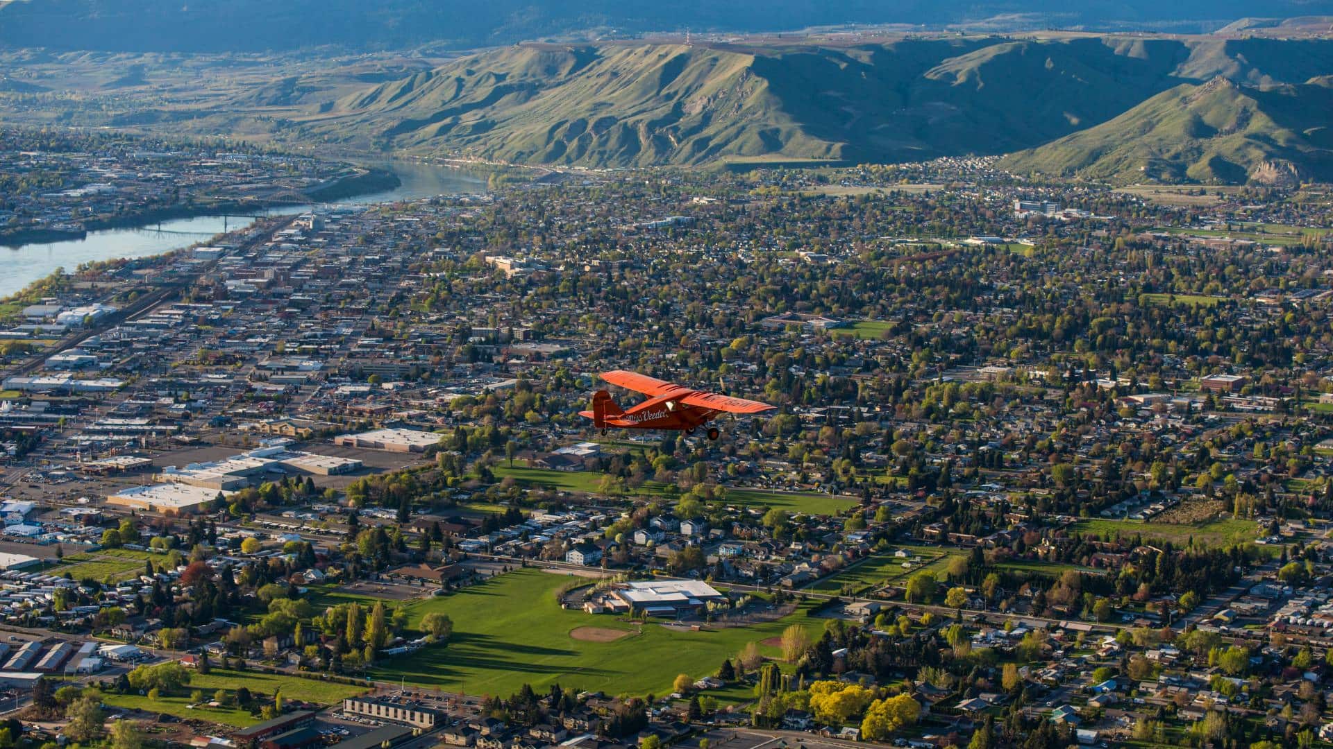 Red airplane flying in the sky over a town with hills in the background