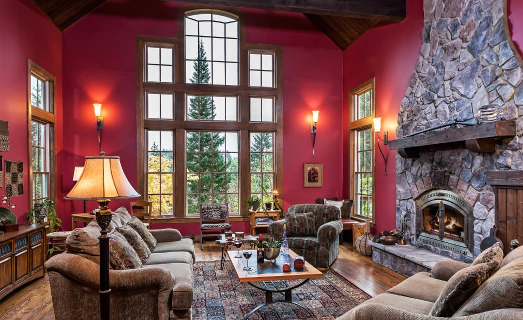 Large great room with dark pink walls, hardwood flooring, large stone fireplace, oversized couch and chairs, and large window with views to the outside