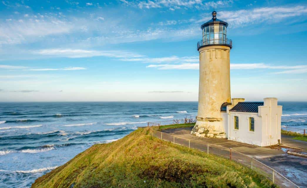 White lighthouse on a hill full of green grass looking over waves on the water