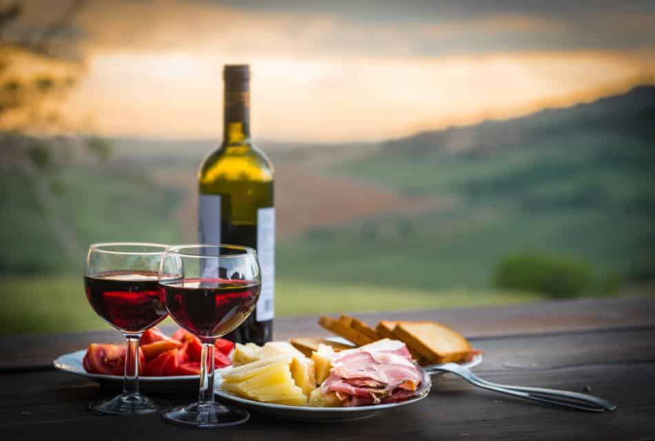 Close up view of two glasses with red wine and white plates filled with meats, cheese, fruit, and bread all sitting on a wooden table