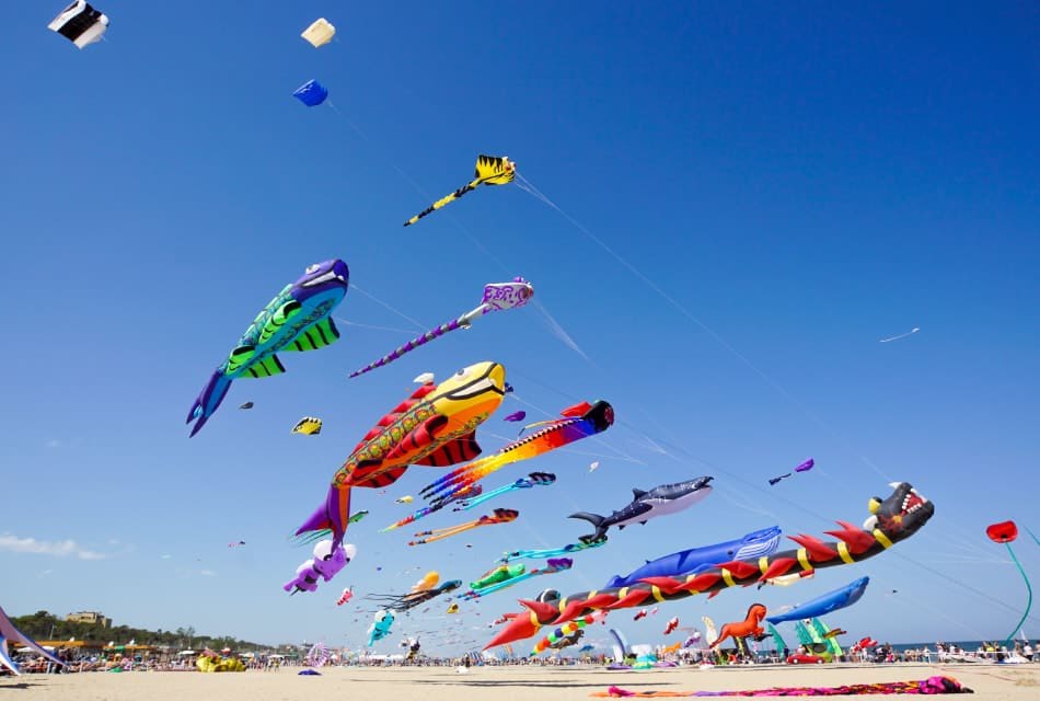 Multiple multicolored kites flying in the sky over a white sandy beach